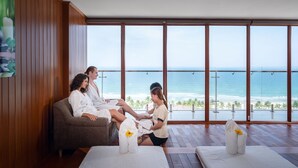 A room with a large window overlooking the ocean, wooden walls, and a seating area with towels and a flower.