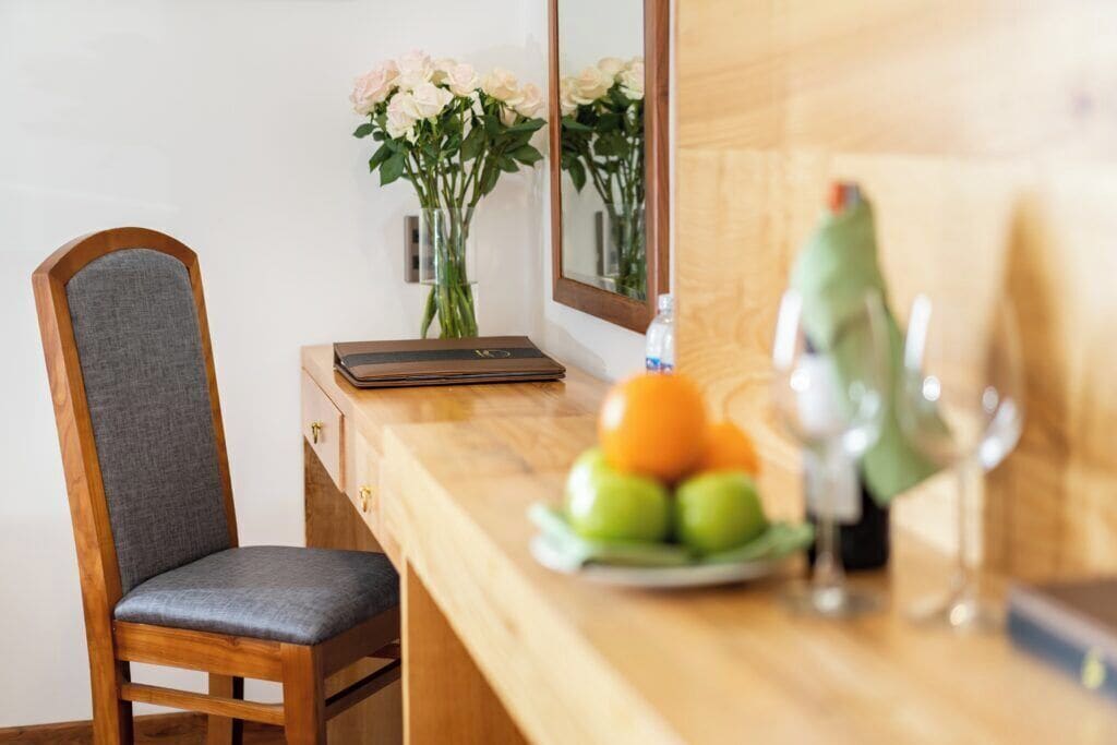 A wooden chair with a grey cushion, a wooden desk with a vase of flowers, a plate of fruit, and a wine glass.