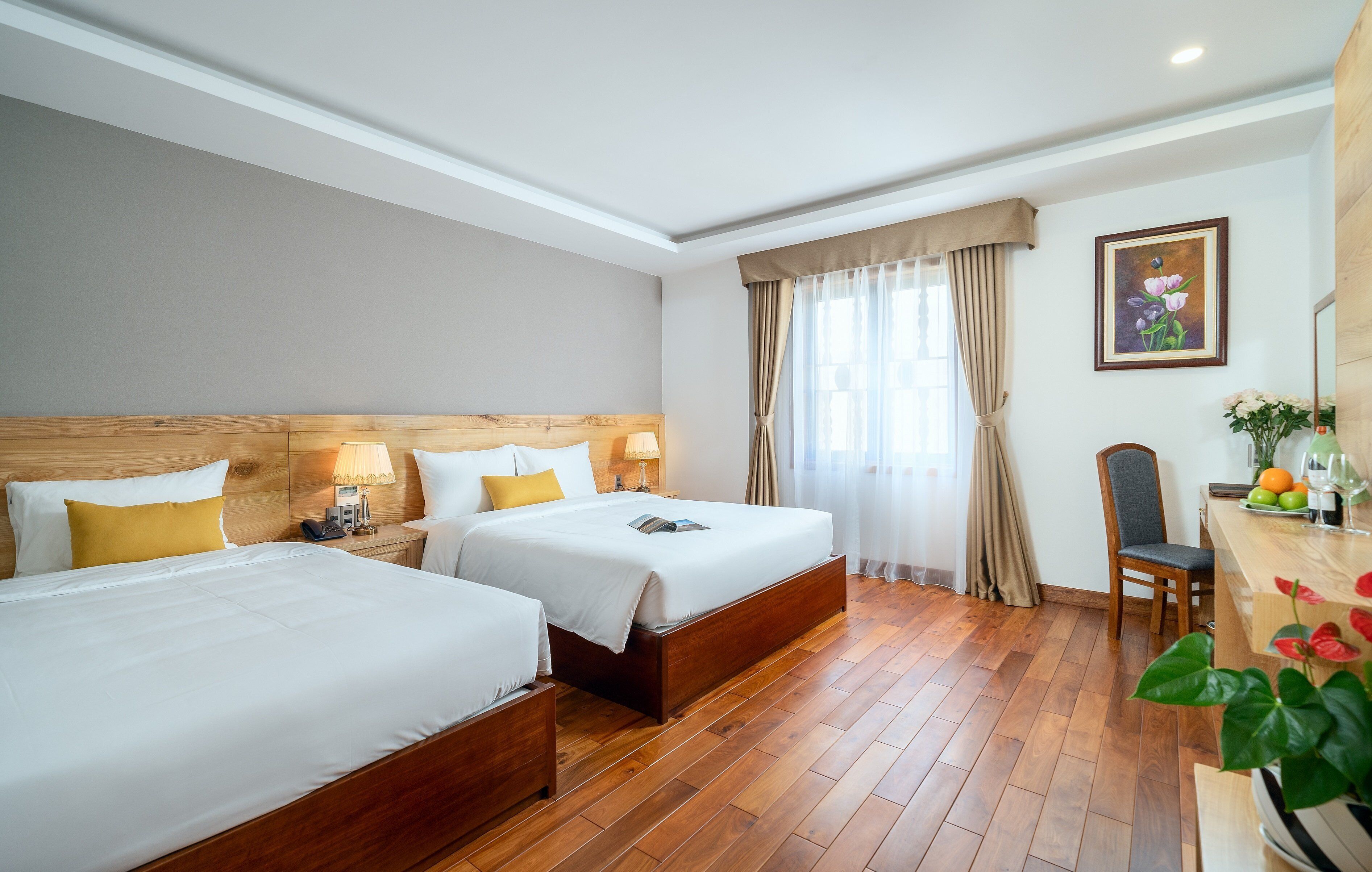 Une chambre d’hôtel avec deux lits, des têtes de lit en bois, un bureau avec une chaise, un vase avec des fleurs et un tableau au mur.
