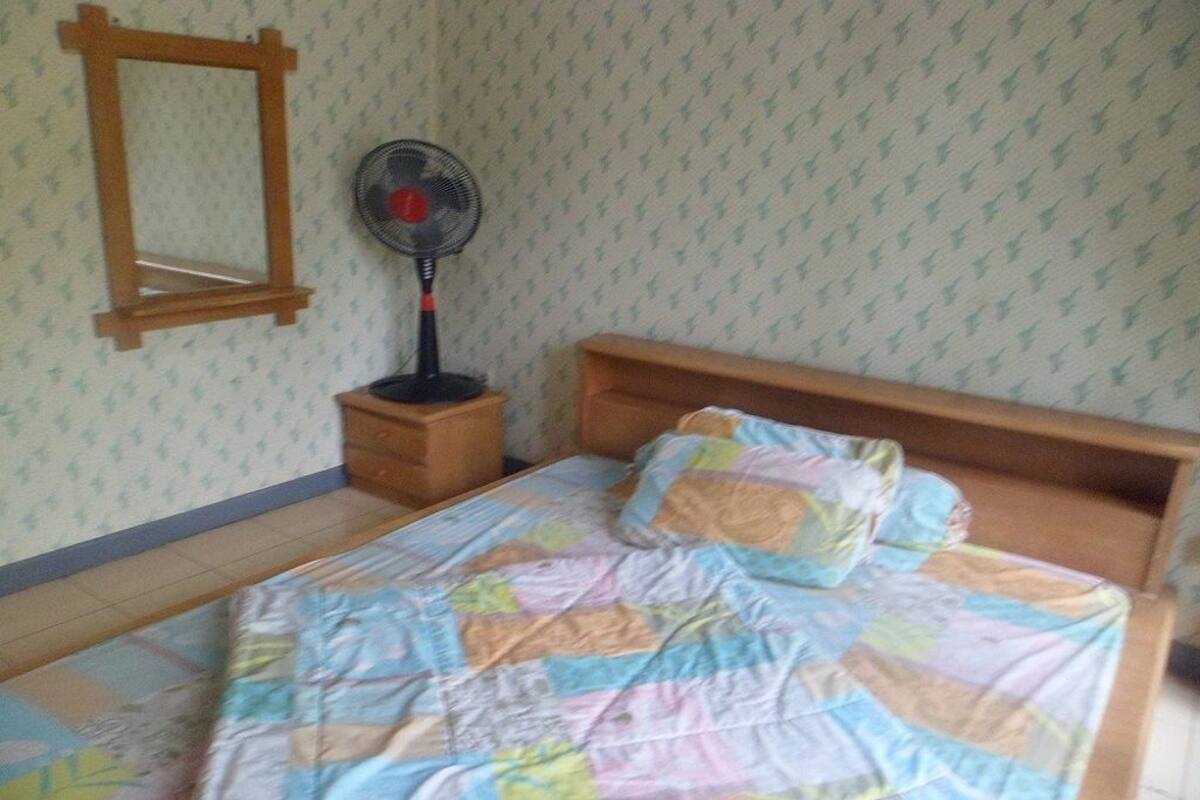 Un dormitorio con una cama, cabecera de madera, mesita de noche con un ventilador y un espejo en la pared.