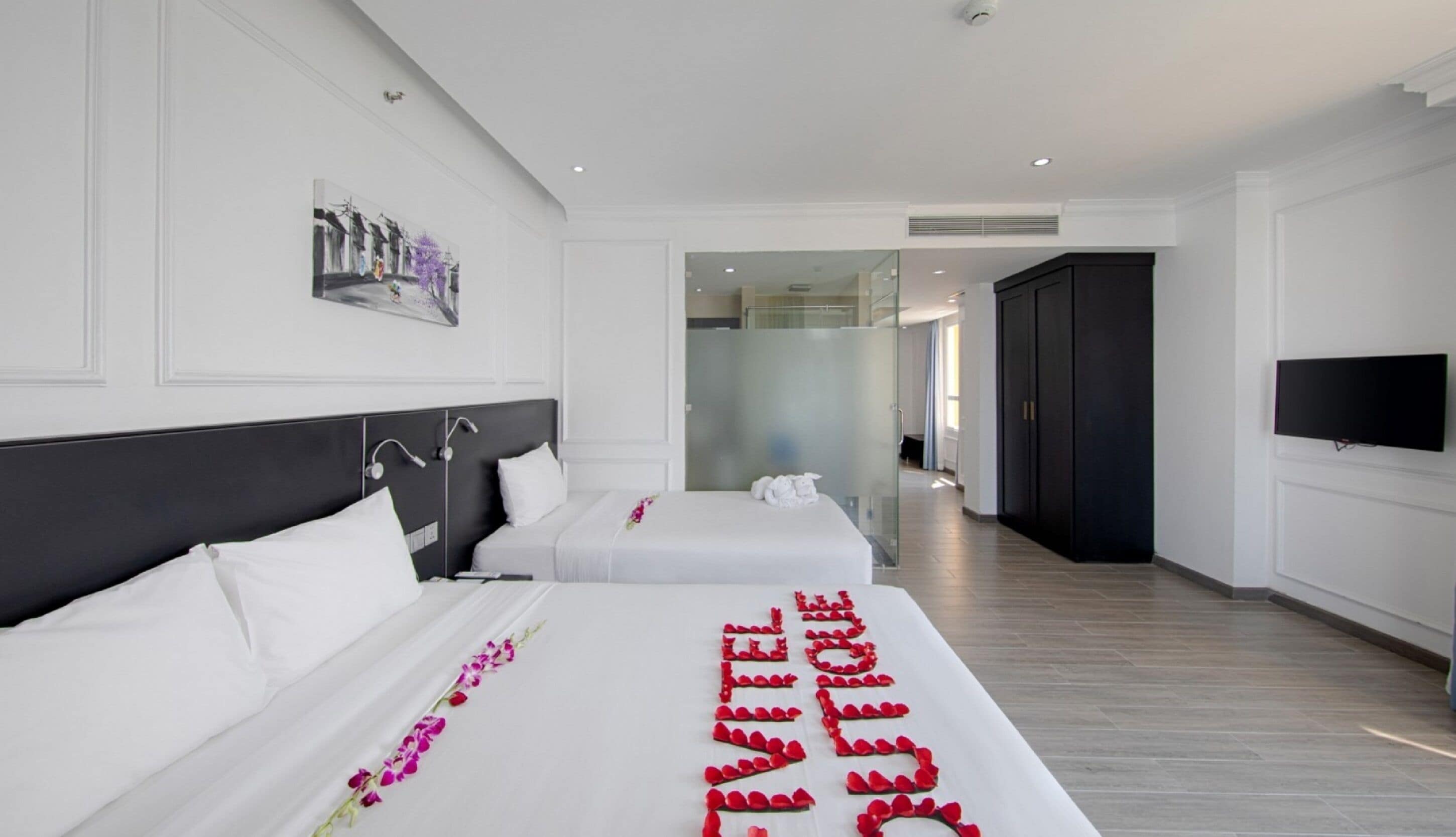 A modern hotel room with two beds, a large mirror, a flat-screen TV, and a hallway leading to a bathroom.