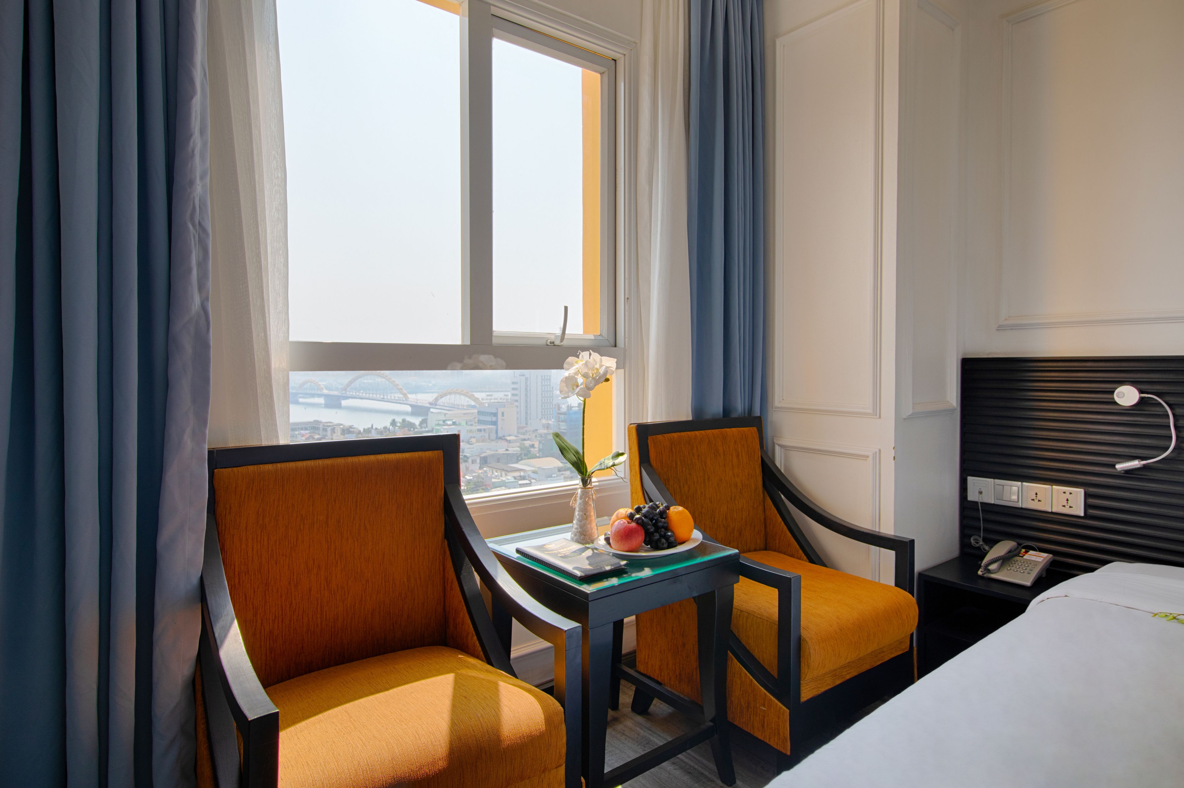 A hotel room with two chairs, a small table with a laptop and fruit, a bed, a nightstand with a phone, and a window with curtains.