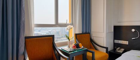 A hotel room with two chairs, a small table with a laptop and fruit, a bed, a nightstand with a phone, and a window with curtains.