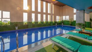 Una piscina interior con baldosas azules cristalinas, ventanas iluminadas por el sol y plantas verdes alrededor del área de la piscina.