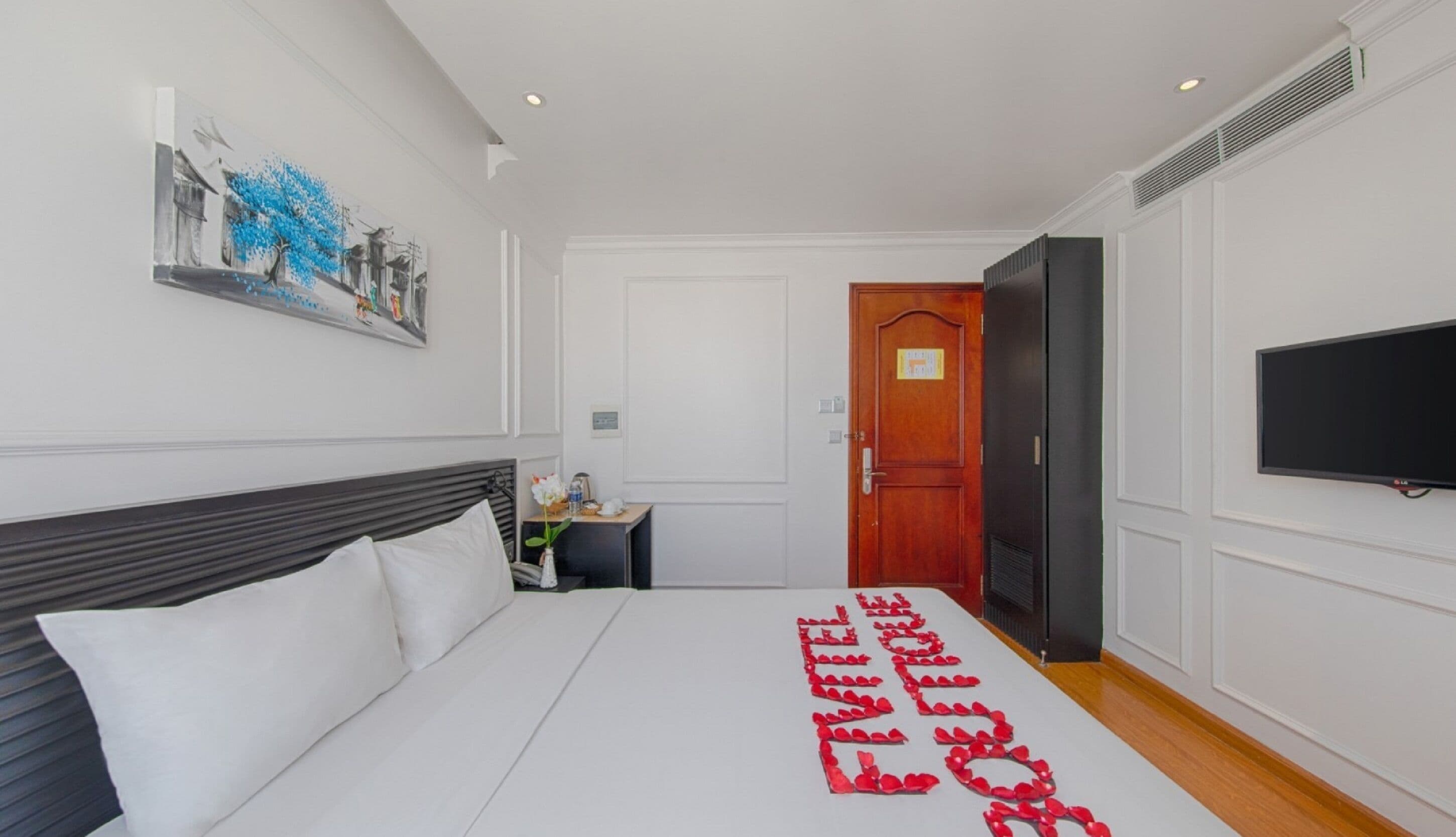 A hotel room with a bed, a TV, a door, and a painting on the wall.