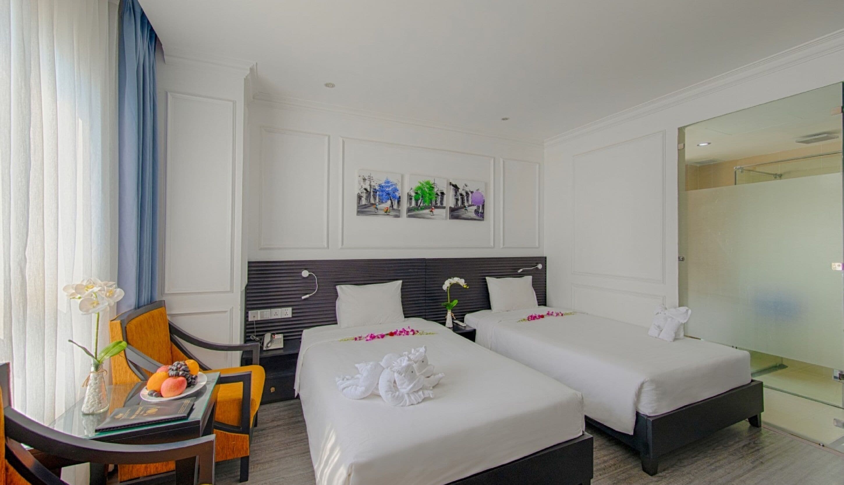 A hotel room with two single beds, a desk with a chair, a vase with flowers, and a wall-mounted artwork.