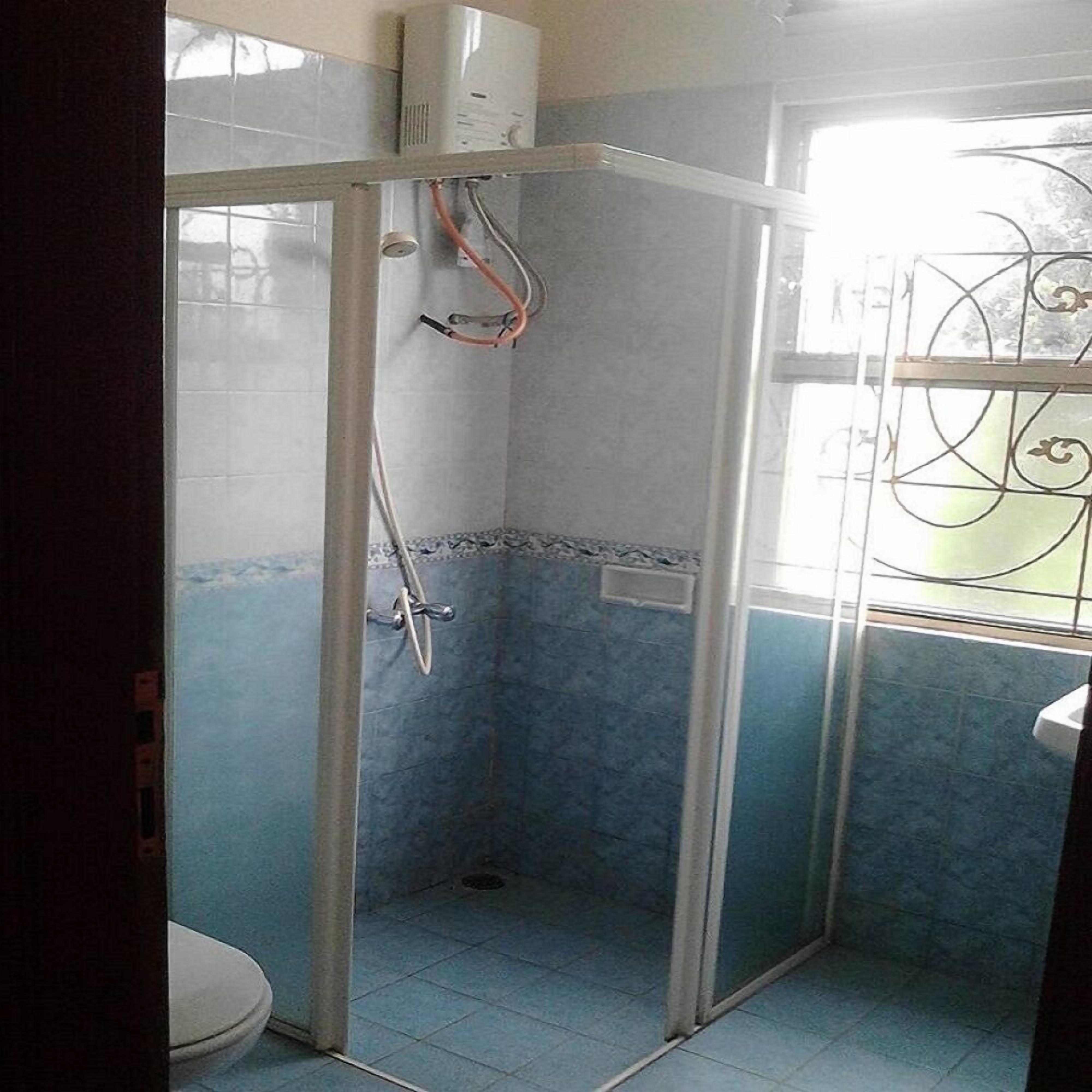 Un espacio de ducha pequeño y cerrado con puerta de vidrio, paredes con azulejos y una ventana con barrotes.
