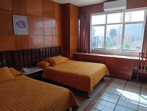 A hotel room with two beds, a desk, a chair, an air conditioning unit, and a window with a view of buildings.