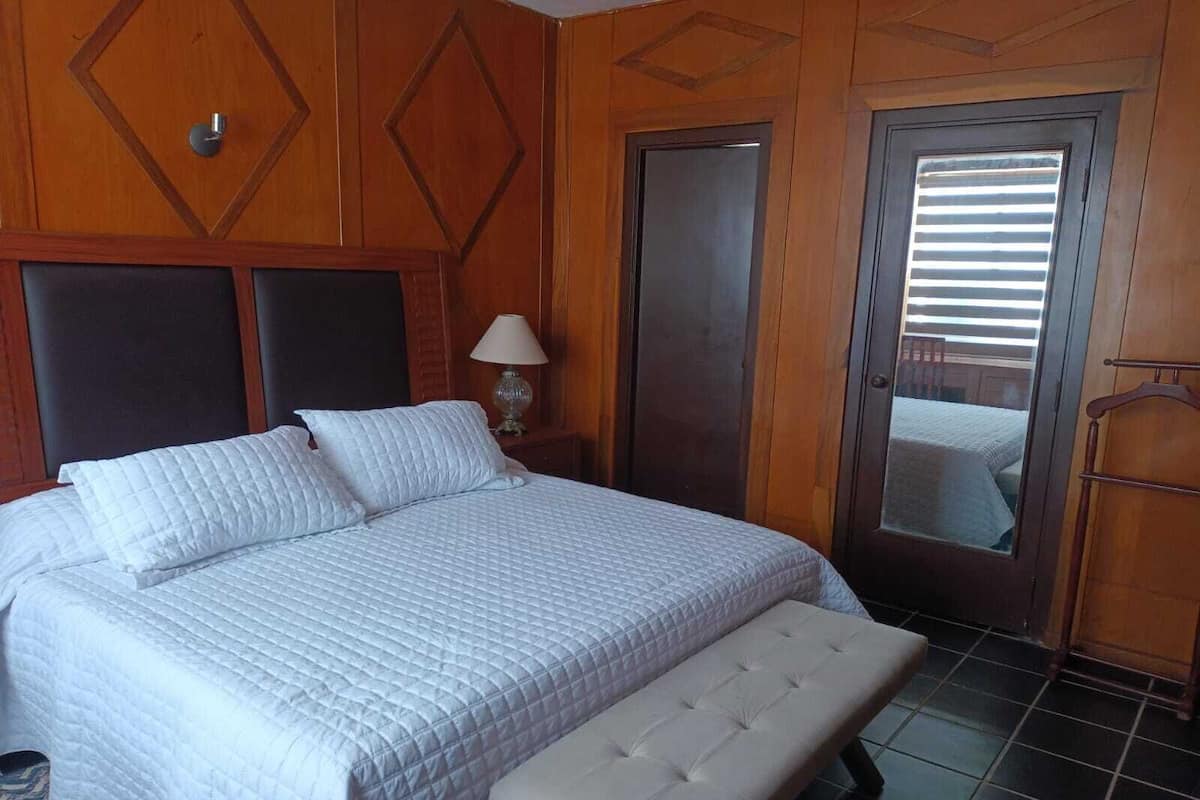 Habitación de hotel con una cama grande, un banco, una mesita de noche con lámpara y una puerta con espejo.