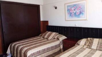 A hotel room with two beds, a dark wooden wardrobe, a framed floral painting on the wall, and a small bedside table.