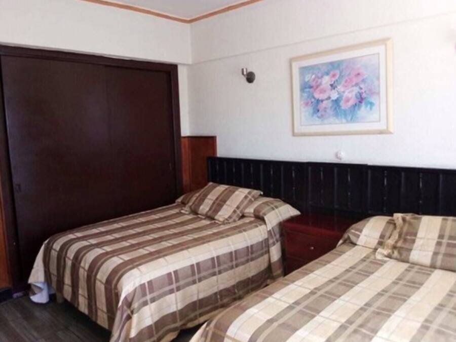 A hotel room with two beds, a dark wooden wardrobe, a framed floral painting on the wall, and a small bedside table.