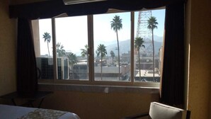 Habitación de hotel con cama, una silla y un amplio ventanal que ofrece vistas a palmeras y edificios.