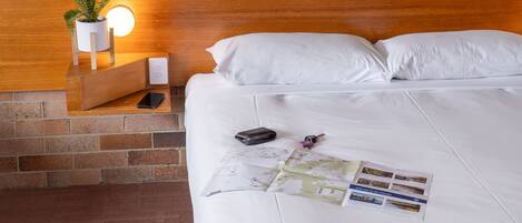 Una cama bien hecha con sábanas blancas, una mesita de noche con una planta en maceta, una lámpara y algunos objetos, incluyendo una billetera y un mapa.