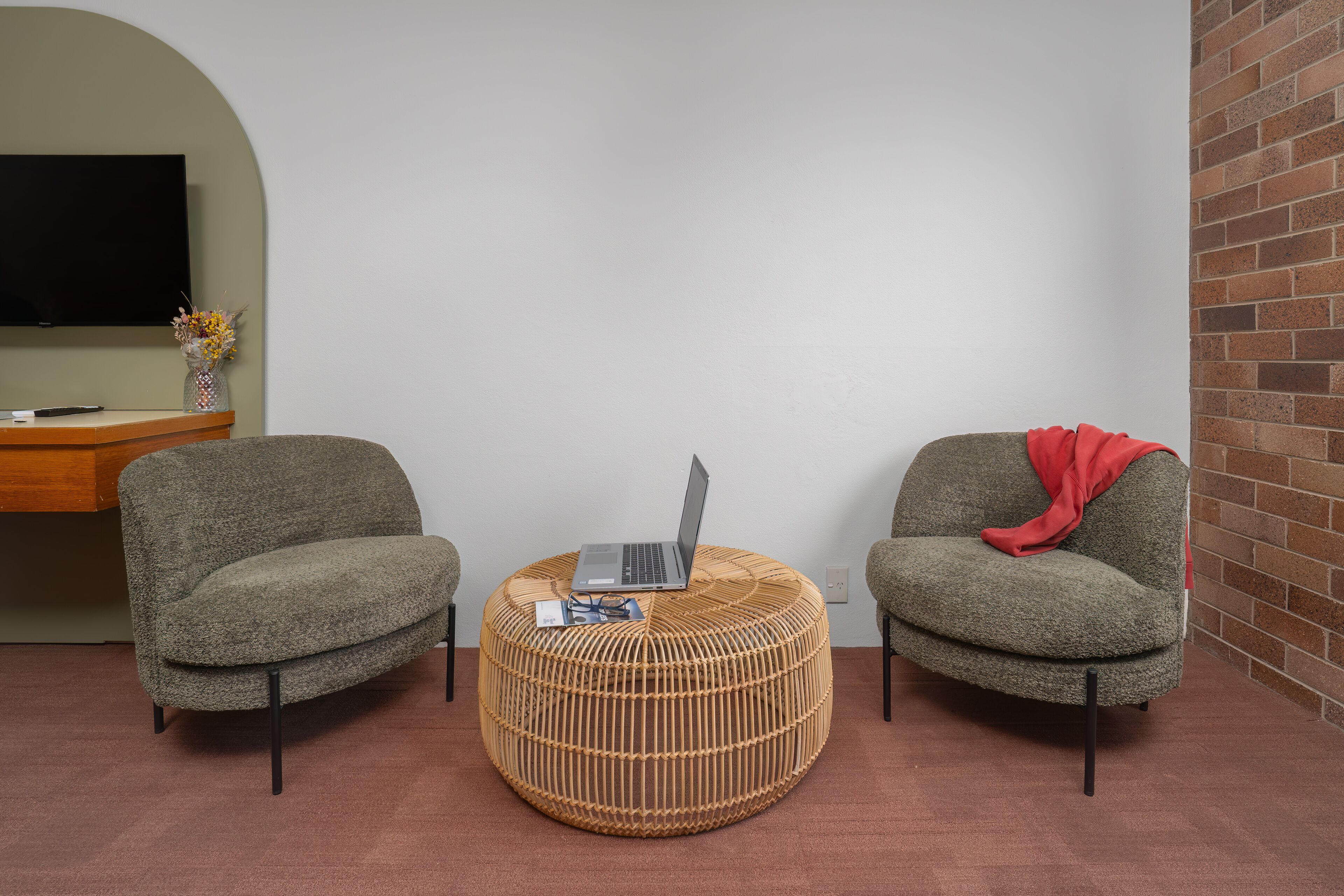 Um quarto com duas poltronas cinzentas, um banquinho redondo de vime com um laptop, e uma televisão na parede.