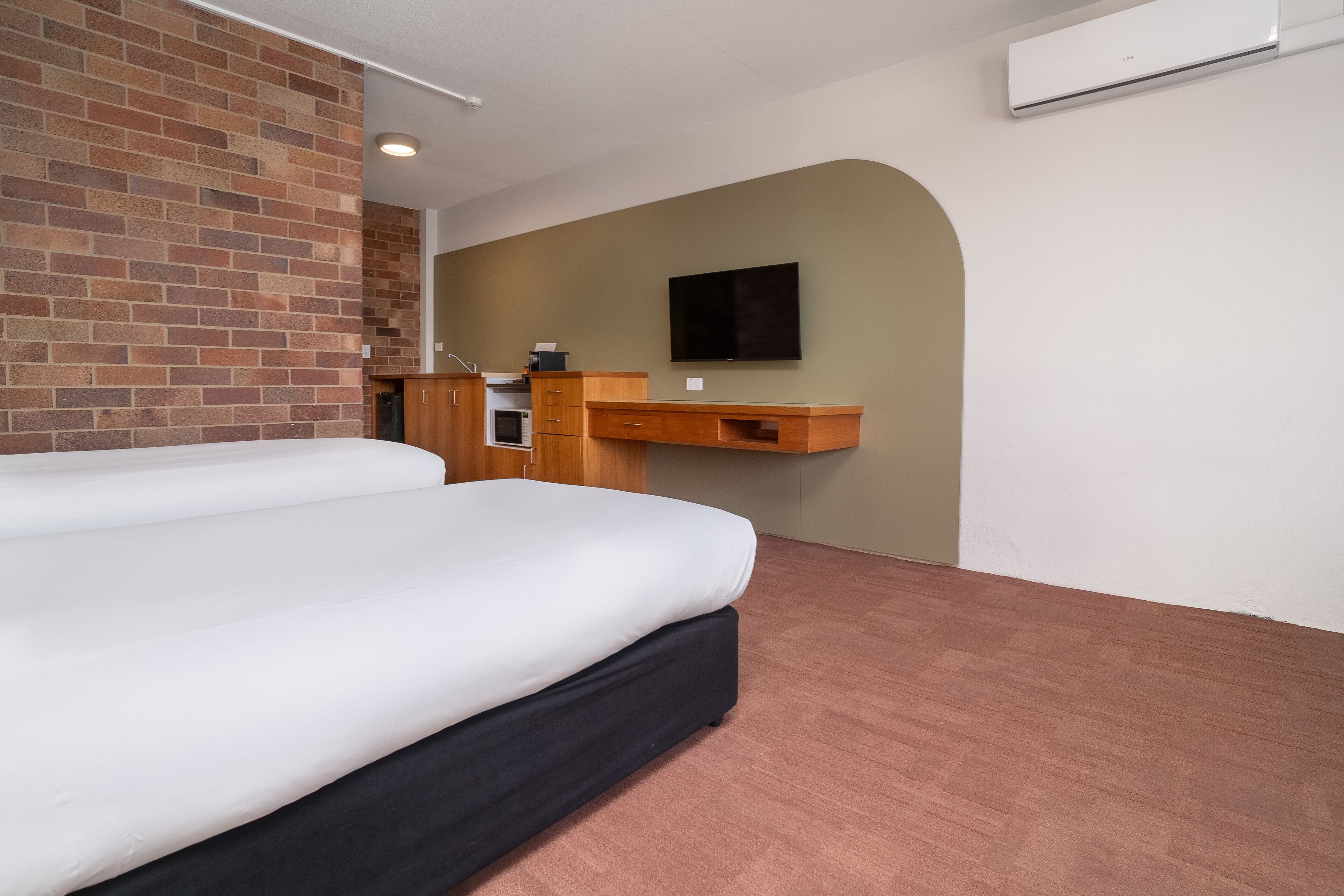 A hotel room with a large bed, a wooden desk, a flat-screen TV, a brick accent wall, and a kitchenette.