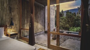 Une chambre avec un mur en bois, un lit, une table de chevet, une lampe et une grande fenêtre donnant sur un jardin et des montagnes.
