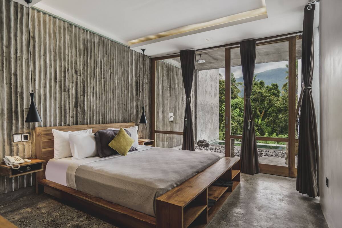 A bedroom with a wooden bed, a nightstand with a phone, and a view of greenery through a glass door.