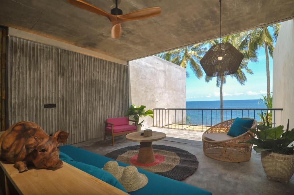 A modern room with a concrete ceiling, a ceiling fan, a wooden table, a pink sofa, a wicker chair, and a view of the ocean.