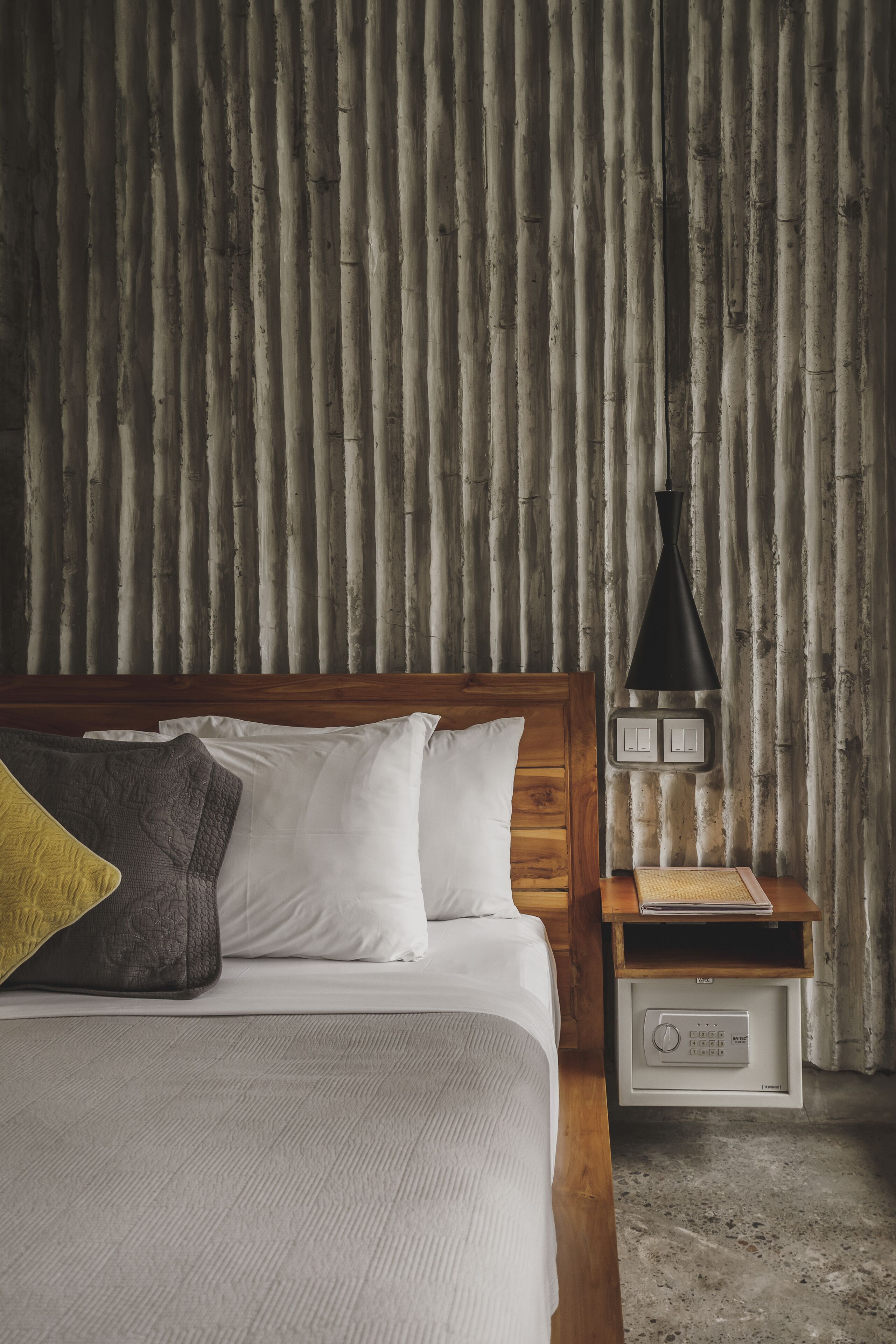 Un dormitorio con cabecera de madera, cama con ropa de cama blanca y gris, mesita de noche con lámpara y pared con listones de madera verticales.