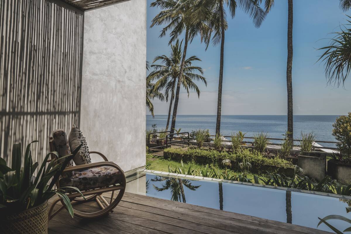 A wooden deck with a wicker chair and a potted plant, overlooking a pool and palm trees with a view of the ocean.