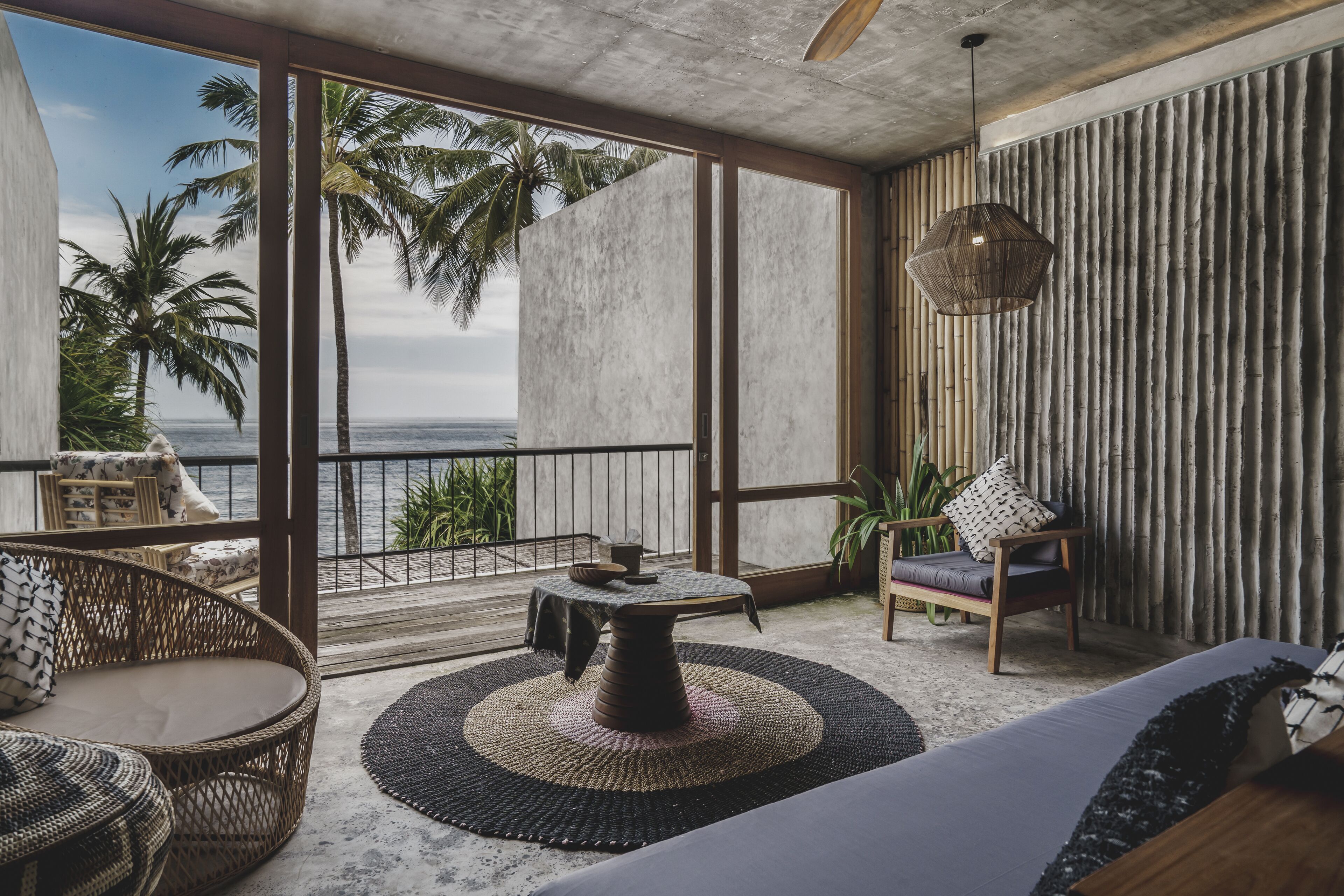 A modern room with a large window overlooking the ocean, featuring wicker furniture, a round table, and a hanging light fixture.