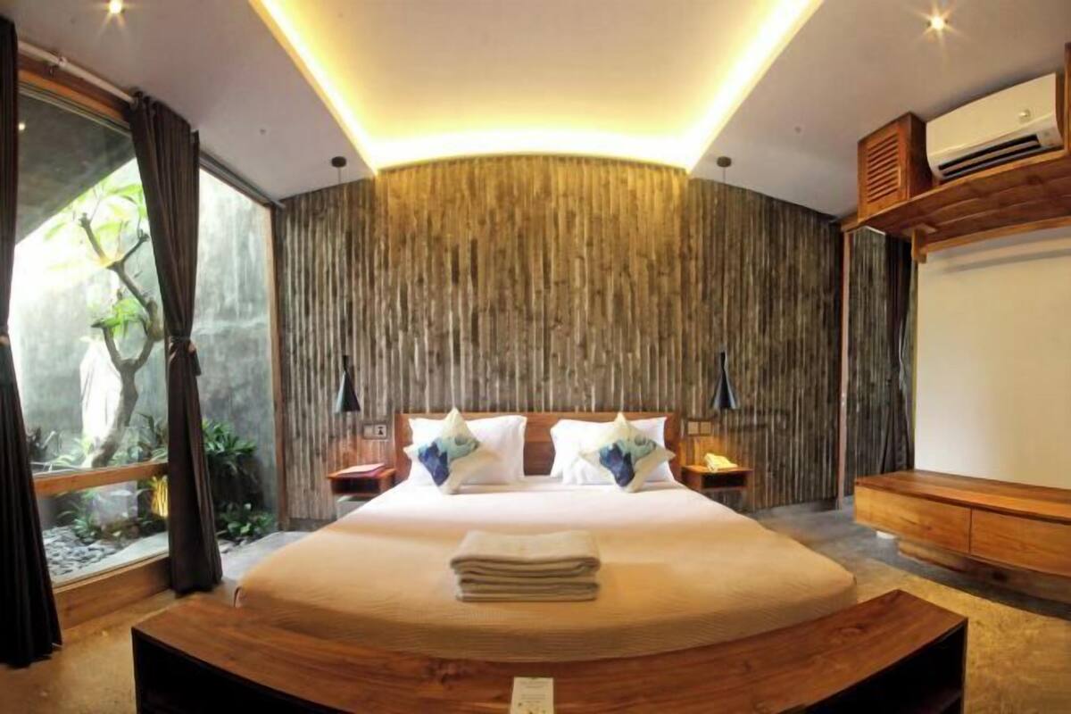 A modern bedroom with a large bed, wooden headboard, and a view of a tree through a window.