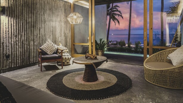 Un salon moderne avec vue sur l’océan au coucher du soleil, comprenant un canapé, une table ronde et un fauteuil en osier.