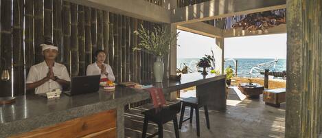 A reception area with a wooden counter, two staff members, a laptop, and a view of the sea through large windows.