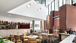 A modern restaurant with a high ceiling, exposed brick walls, and a variety of seating arrangements including round tables and booths. - Veriu Green Square (Alexandria)