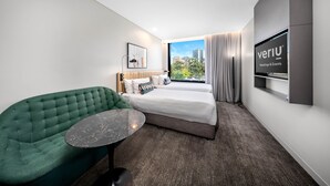 A modern hotel room with a green sofa, a bed with white linens, a flat-screen TV, and a city view. - Veriu Green Square (Alexandria)
