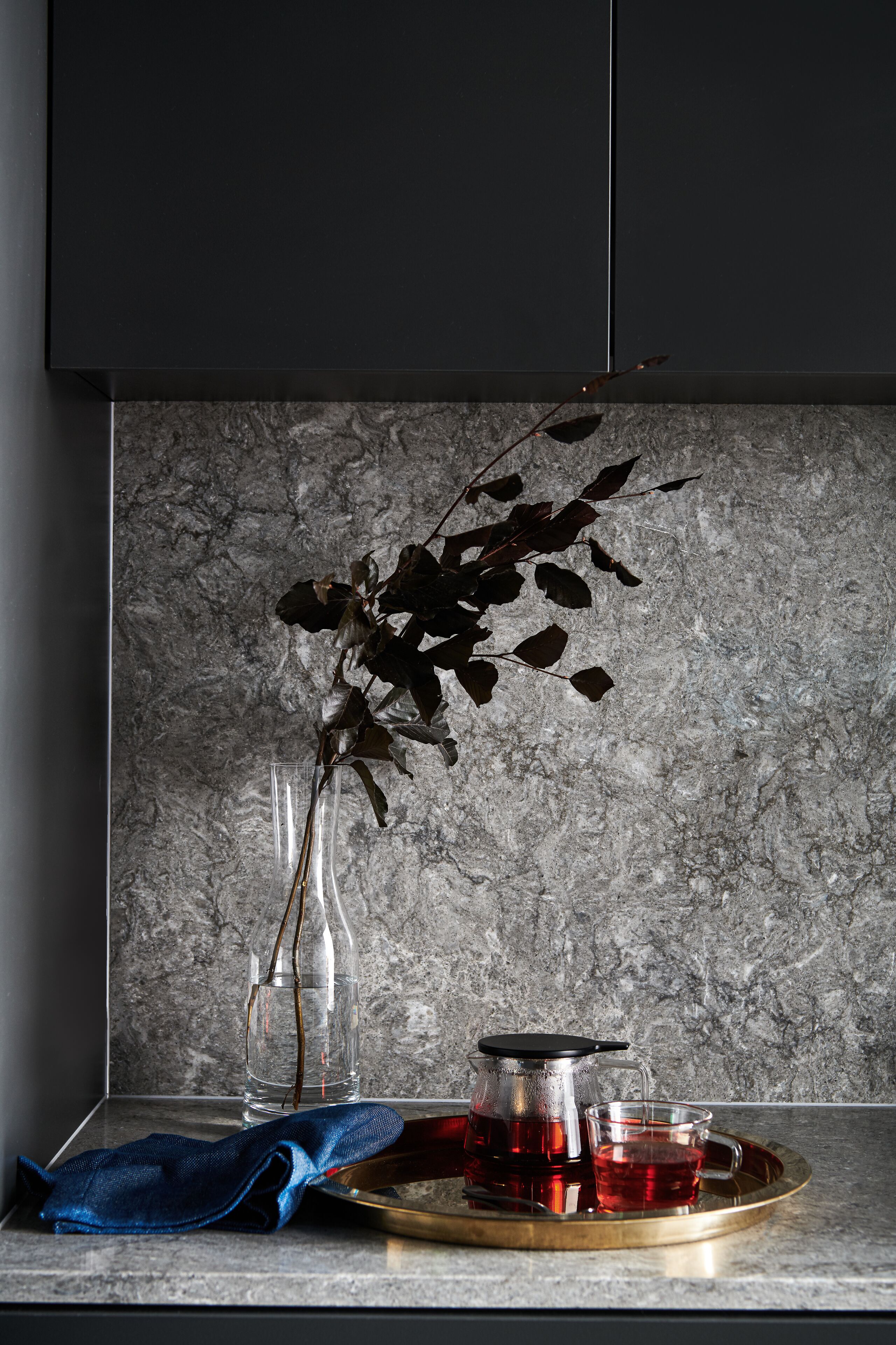 A kitchen counter with a tea set, a clear glass vase with a dried plant, and a blue cloth.