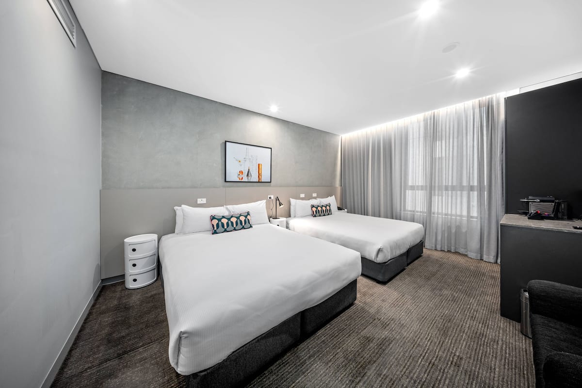 Et moderne hotelværelse med to senge, et skrivebord, en stol og et stort vindue med gardiner.