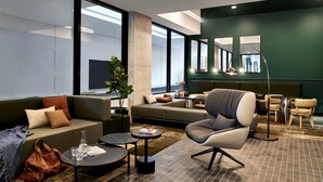A modern hotel lobby with a sofa, armchair, and small tables, large windows, and a green accent wall. - Veriu Green Square (Alexandria)