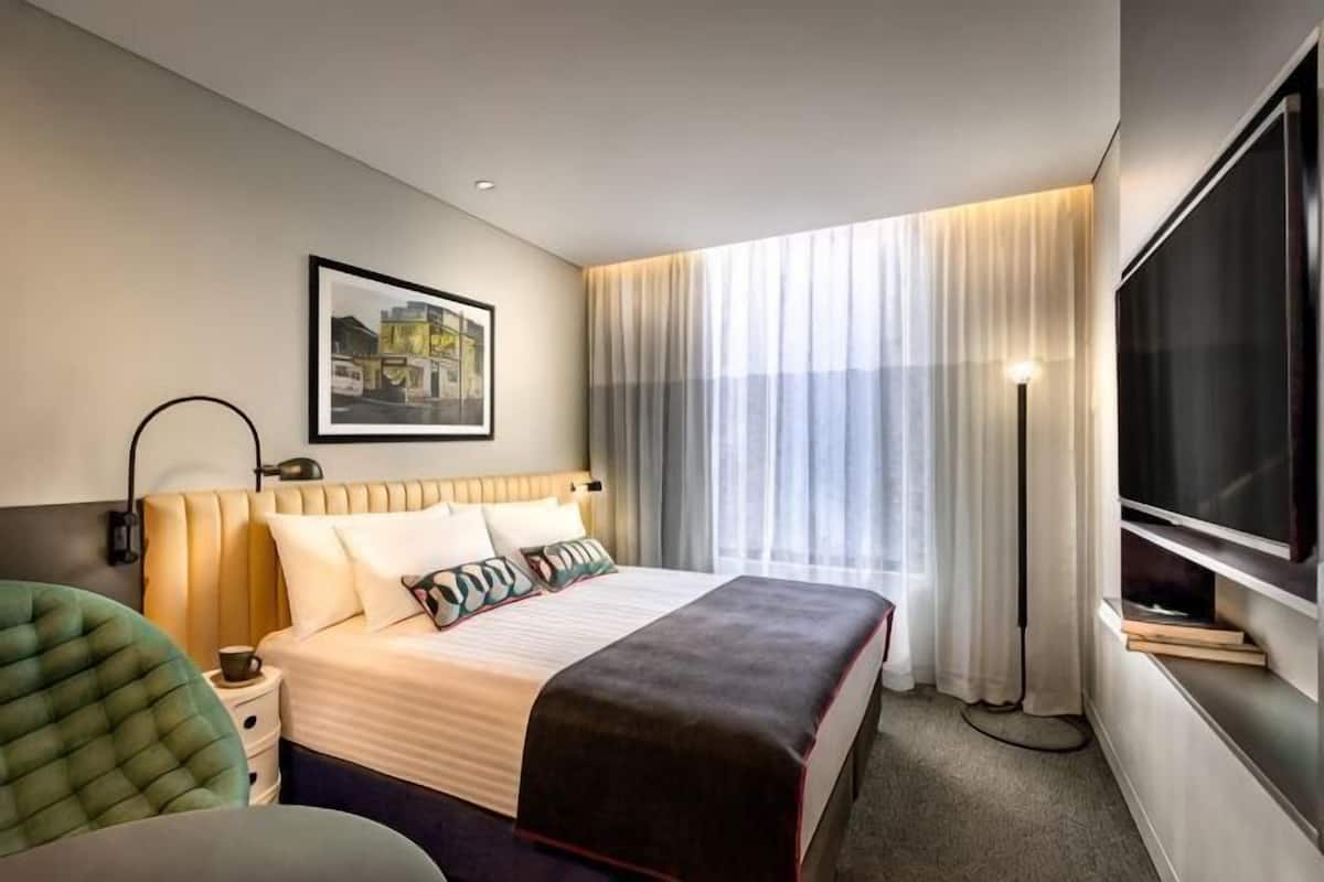 Et hotelværelse med seng, skrivebord, stol, fjernsyn og et vindue med gardiner.