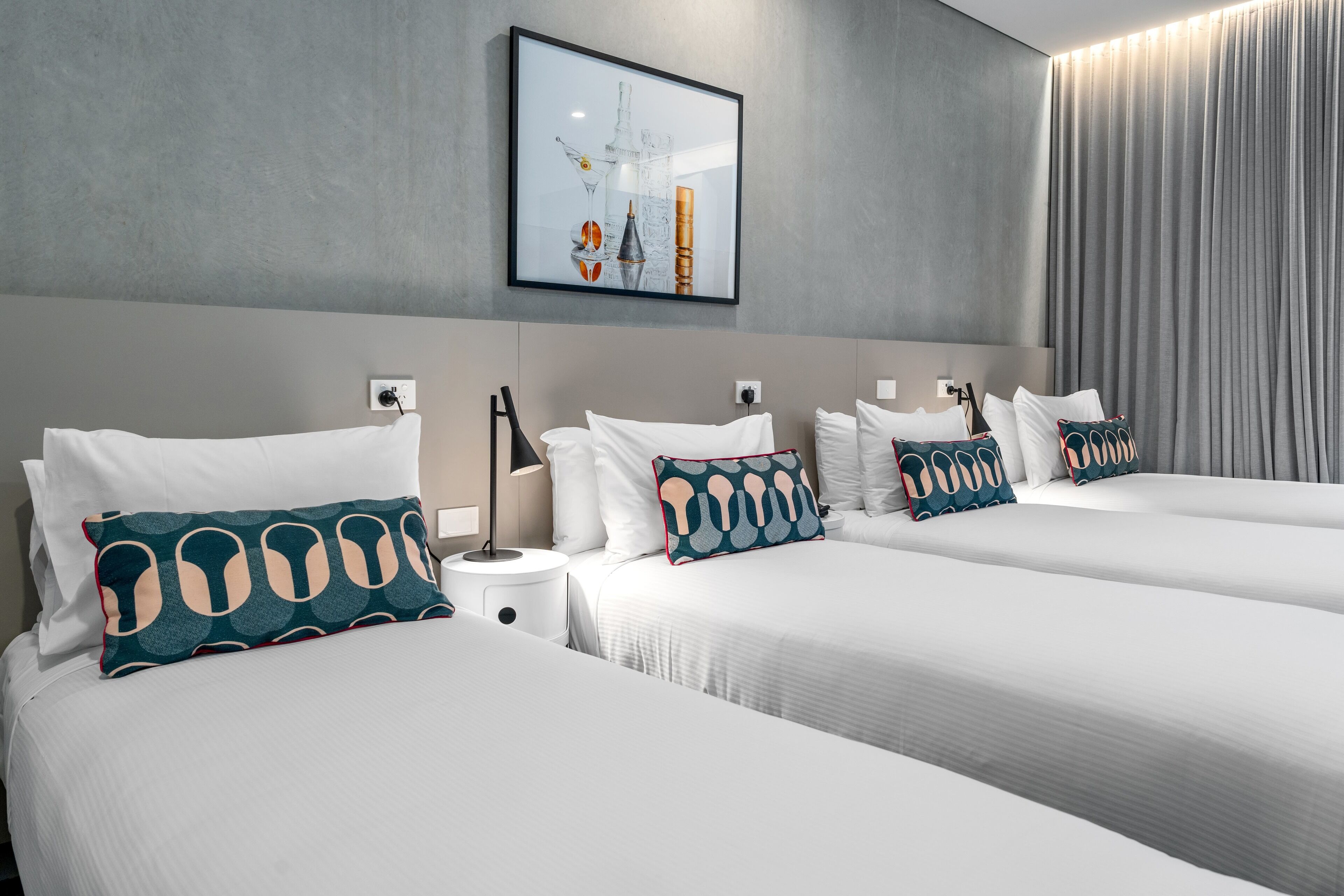 A modern hotel room with two beds, a large window with curtains, a wall-mounted artwork, and a bedside table with a lamp.