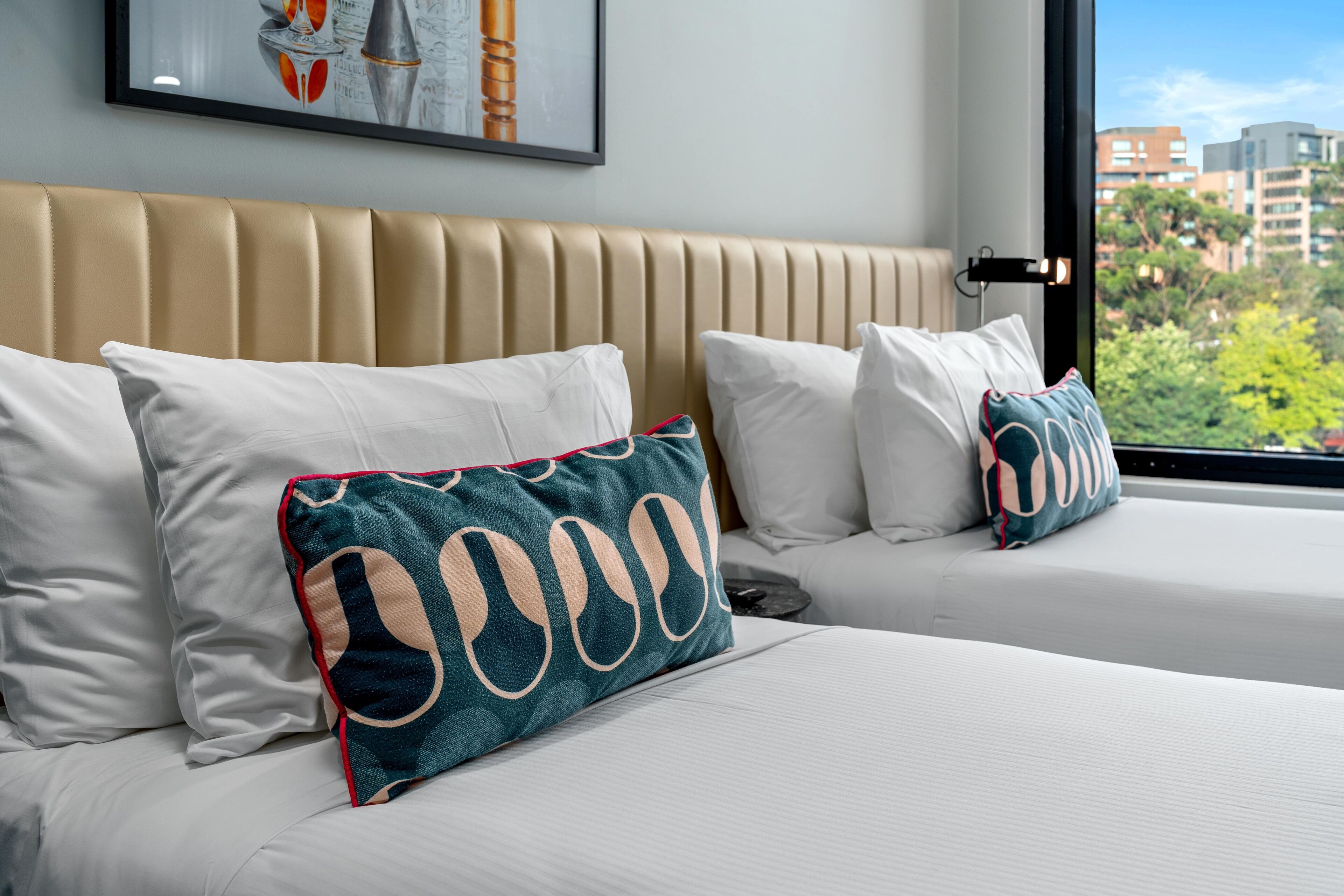 A hotel room with two beds, beige headboard, white bedding, and a city view through a large window.