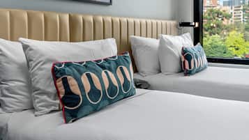 A hotel room with two beds, beige headboard, white bedding, and a city view through a large window.