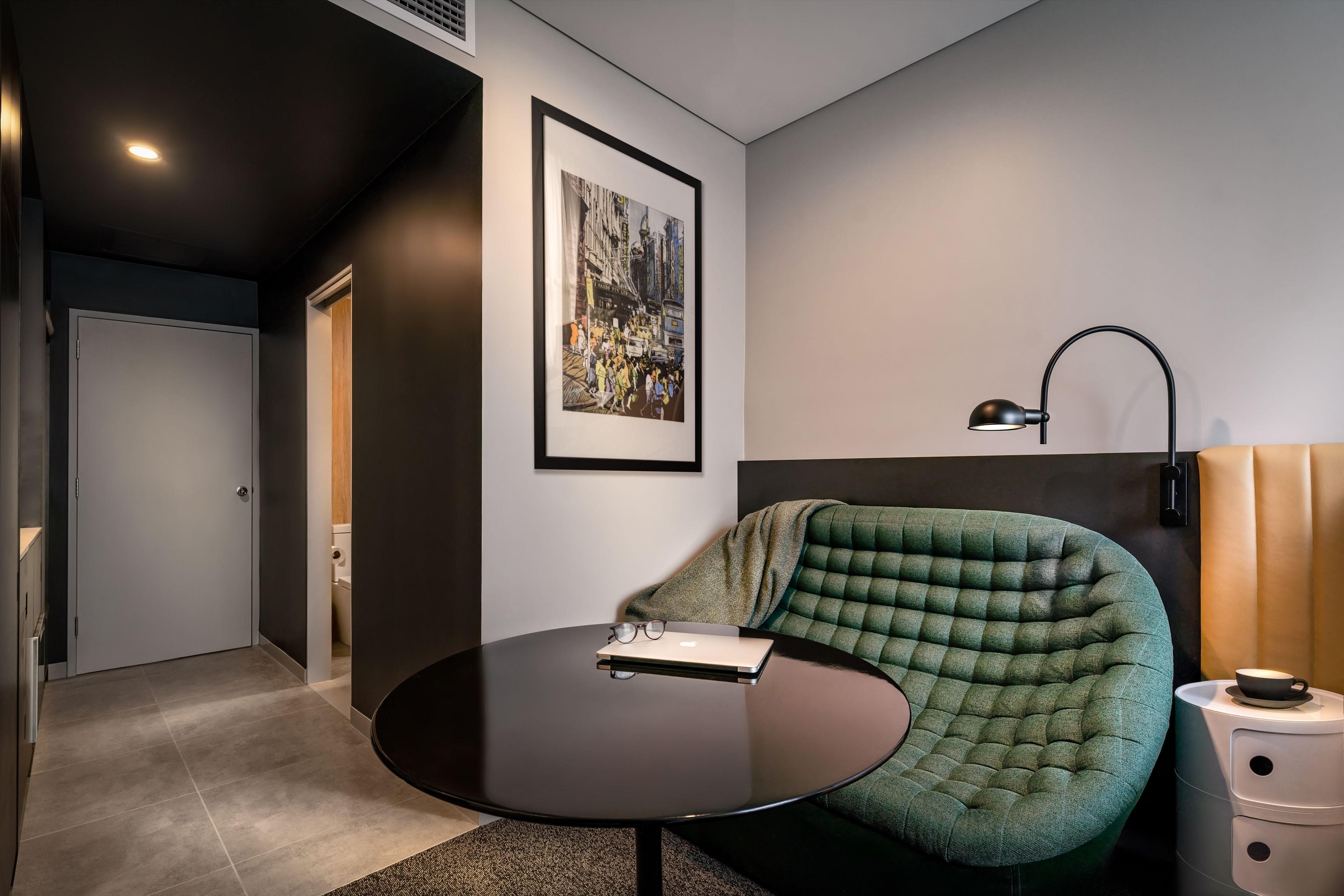 Une chambre moderne avec une table ronde, un fauteuil vert à motifs, un tableau représentant un paysage urbain encadré et une lampe de chevet.