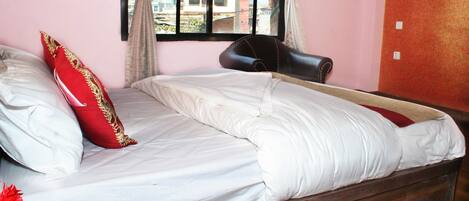Una cama individual con ropa de cama blanca y una almohada roja, un cabecero de madera, una ventana con cortinas y vistas a edificios.
