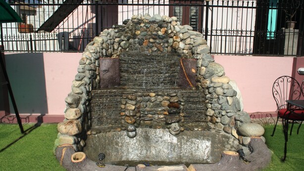 A stone water feature with a cascading design, surrounded by a stone wall and a metal fence. - Sitapaila Homestay and Apartment (Nagarjun)