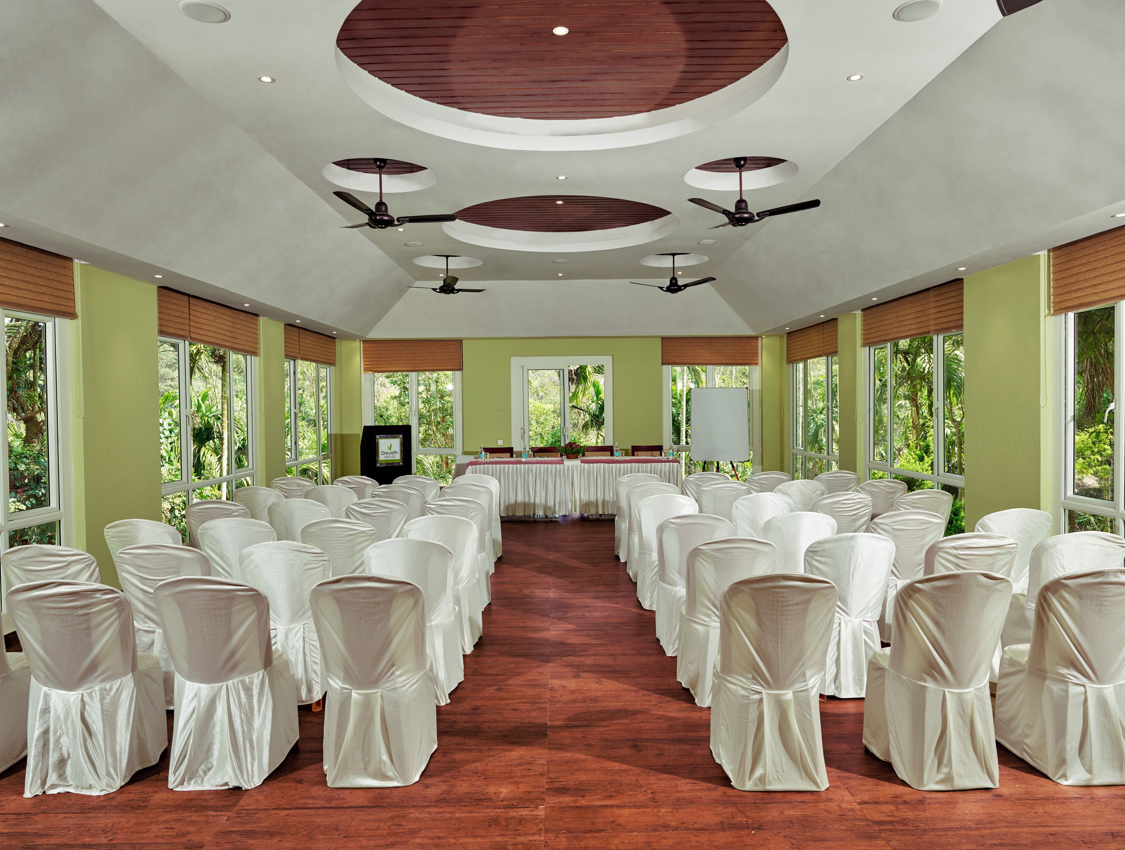 Un salón de conferencias con filas de sillas blancas, ventiladores de techo y amplias ventanas con vista a la vegetación.