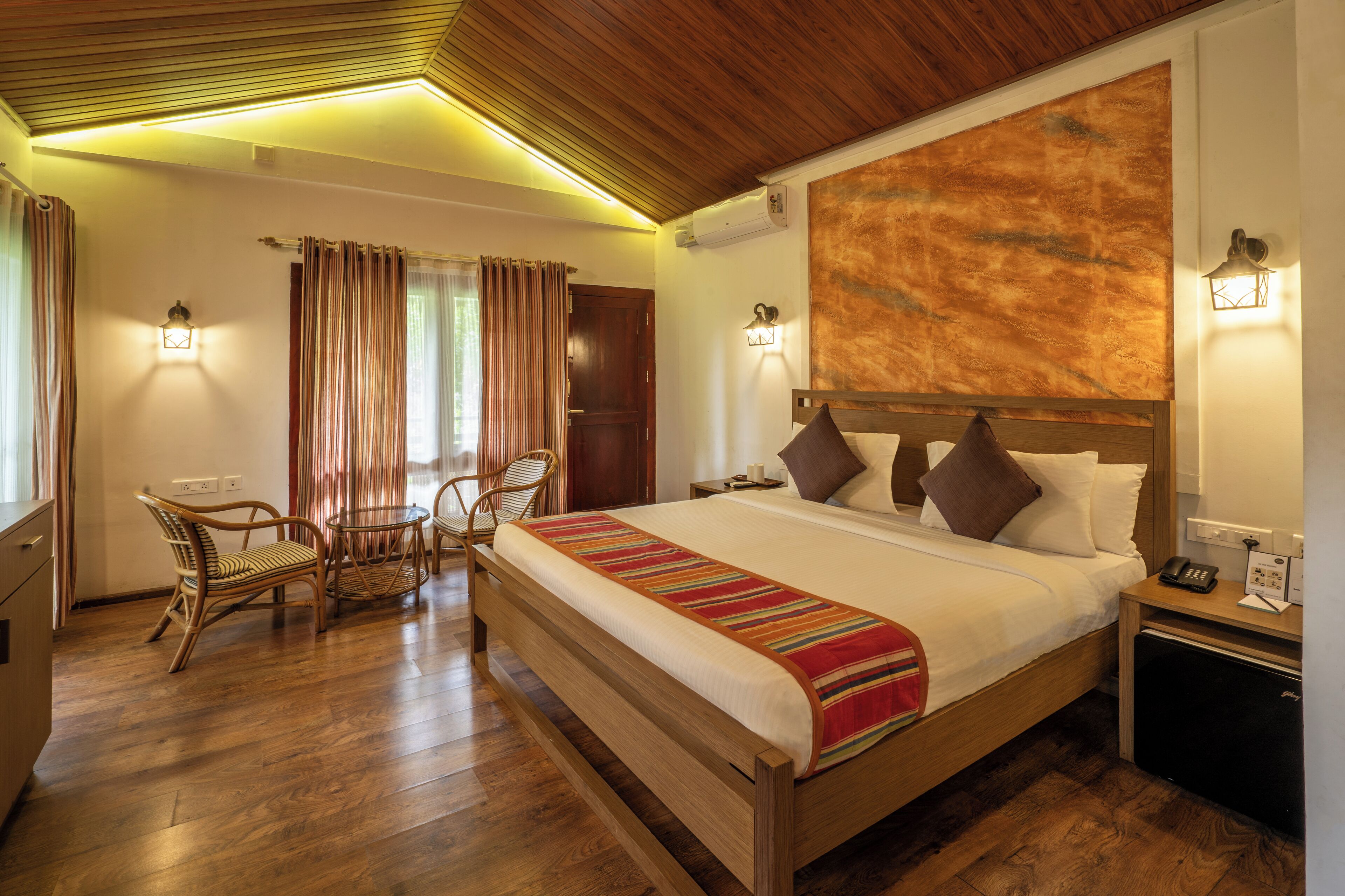 Une chambre d’hôtel avec un lit en bois, une petite table, des chaises en osier et un bureau avec un téléphone et une lampe.
