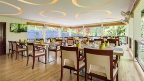 A spacious dining area with multiple tables and chairs, large windows offering a view of greenery, and a modern ceiling design. - The Woods Resorts Wayanad (Sulthan Bathery)