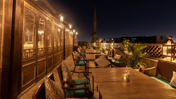 An outdoor dining area with wooden tables and chairs, a view of a tall tower, and a wooden fence. - Le Riad Hotel de Charme (Cairo)