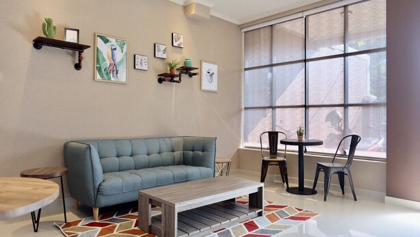 A modern living room with a grey sofa, a wooden coffee table, and two chairs. There are wall-mounted shelves with decorative items and a large window with blinds. - Style50 homestay (Surabaya)