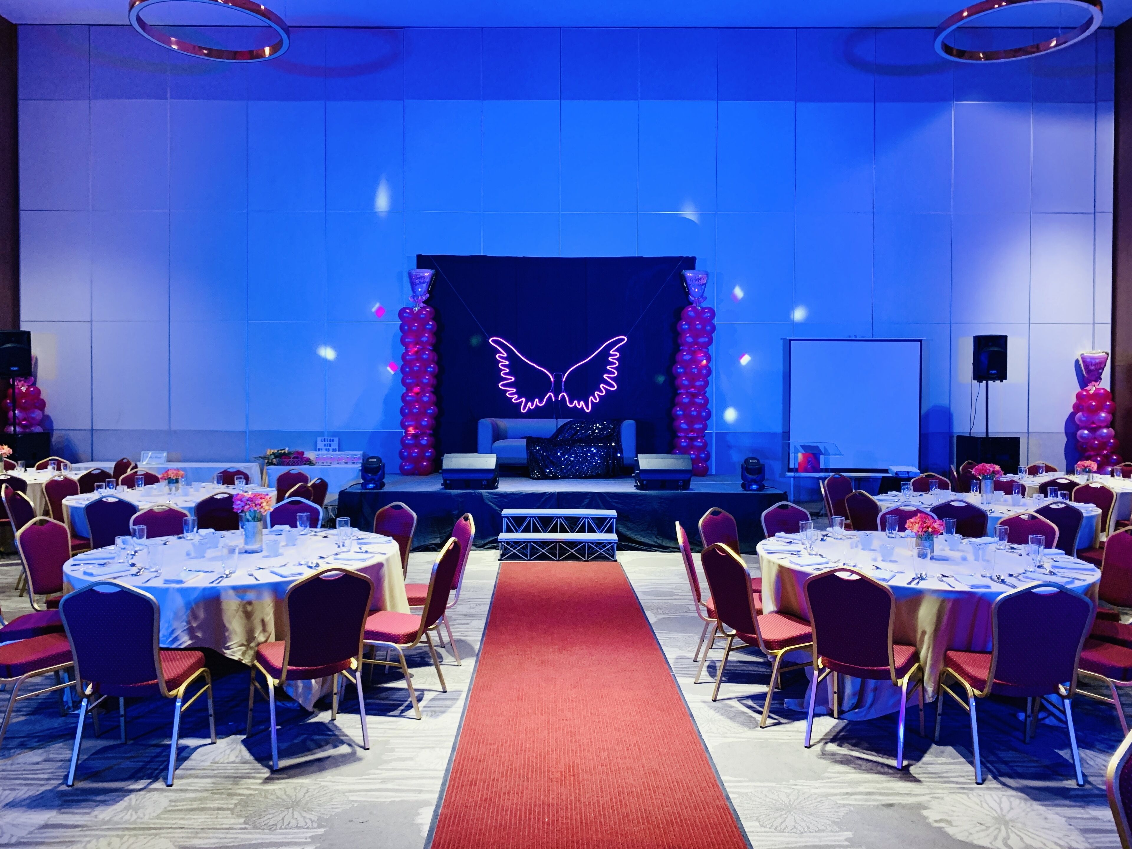 A conference room with round tables, red chairs, and a stage with a neon wing design.