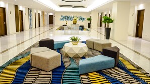 Um lobby de hotel moderno com carpete estampado em padrão geométrico, uma área de estar central com pufes e uma grande pintura abstrata na parede.