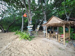 A rustic wooden structure with a thatched roof, surrounded by a sandy area and dense foliage.