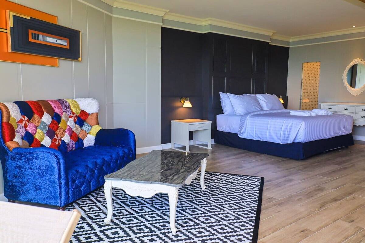 Habitación de hotel con sofá azul, cama con ropa blanca, mesita de noche pequeña y blanca, mesa de centro de mármol y alfombra estampada.