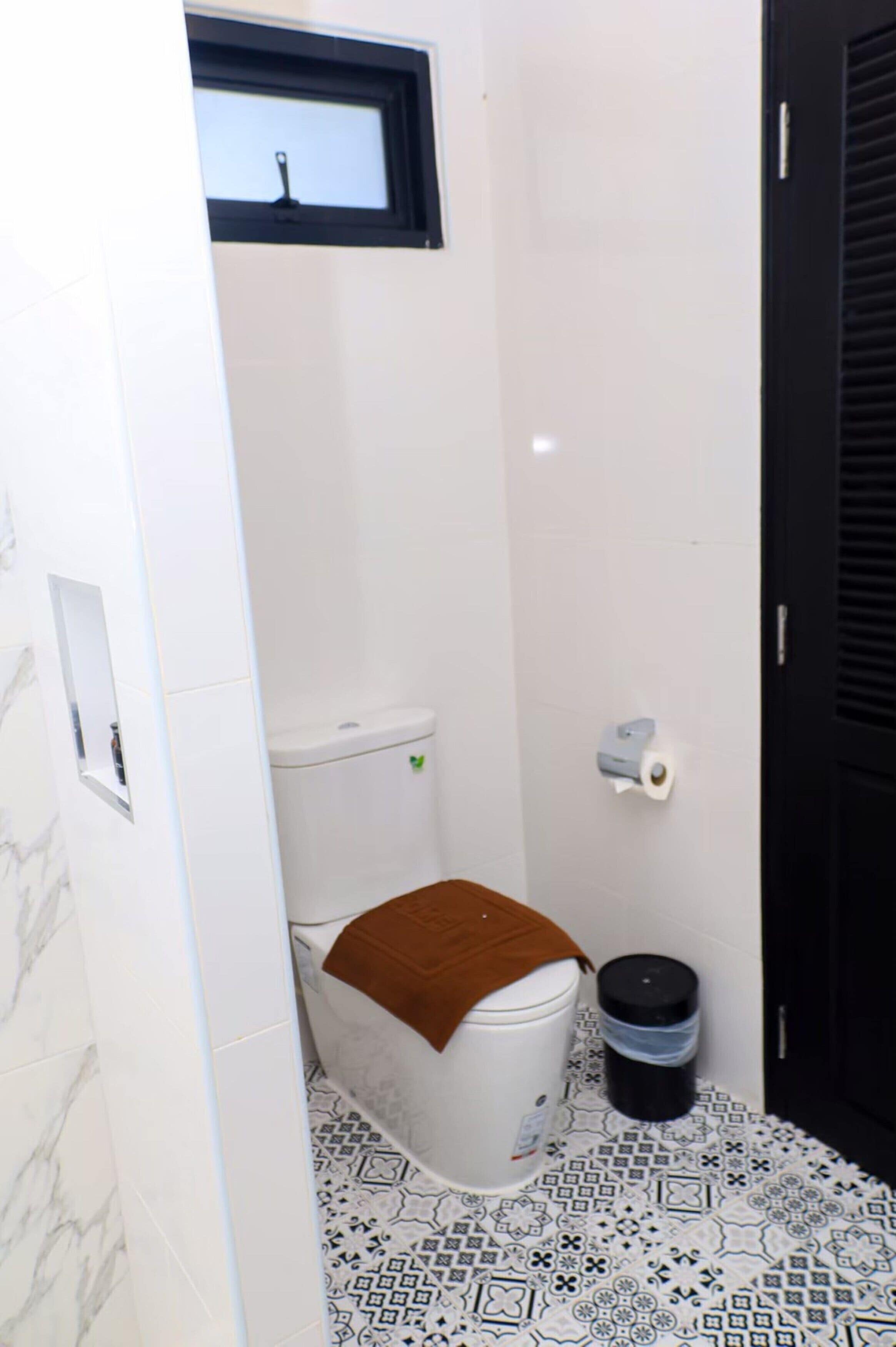 A small bathroom with a toilet, a trash bin, and a window above the toilet.